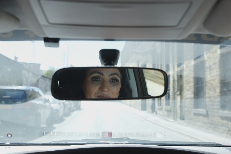 A point-of-view photo taken from inside a taxi, looking out through the front windscreen at a street scene ahead. The dark dashboard runs along the bottom of the frame. In the center, the rear-view mirror clearly captures a close-up reflection of the taxi driver—a South Asian woman wearing a long scarf—looking steadily ahead.