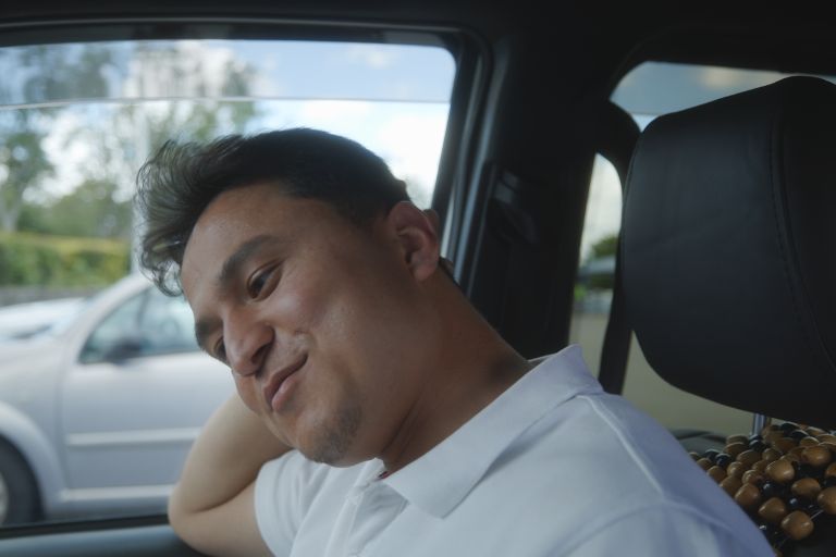 A taxi driver sat in his taxi wearing a white tshirt with a collar, with his head to one side and a cheeky smile on his face.