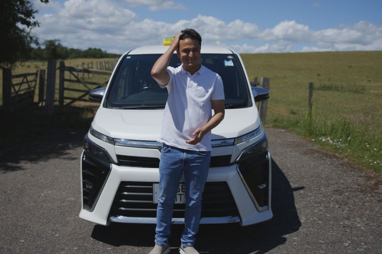 A taxi driver wearing a white polo shirt, blue jeans and white trainers, stands infront of his white taxi that is stationary. Behind him is a field, with blue sky and scattered clouds.