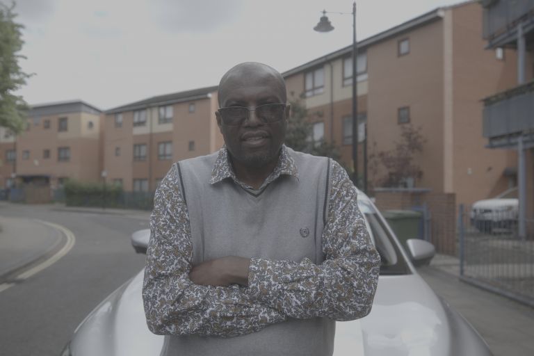 A black taxi driver wearing shades, a grey pullover, patterned shirt is stood infront of his silver taxi with his arms crossed. Behind him is a street lined with modern blocks of flats.