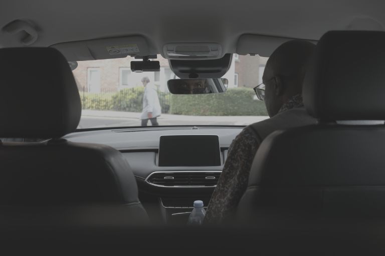 A taxi driver is sat in his taxi looking out through the front window where a pedestrian is crossing the road.