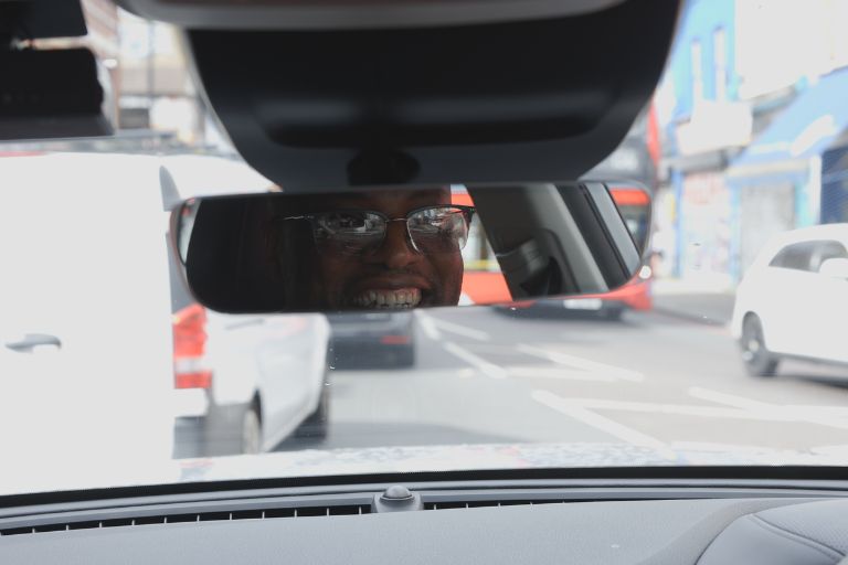 A taxi driver is grinning, his reflection caught in the rearview mirror of his car. He is wearing glasses, and the view infront of him includes lots of white vans parked in an urban street.