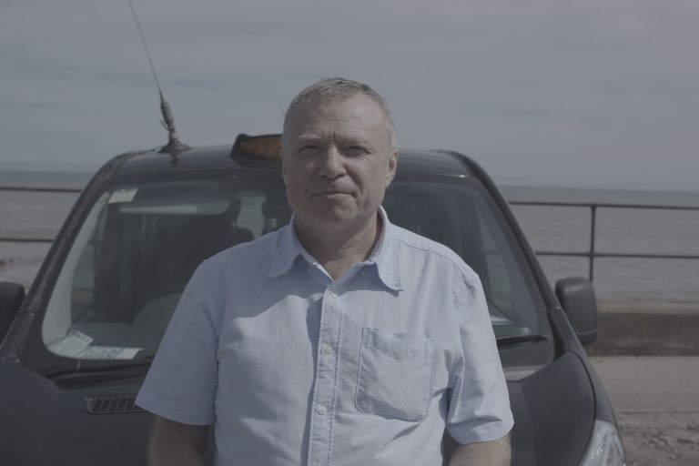 A taxi driver stands in front of his car with a slight smile on his face. He is wearing a light blue shirt, the taxi is black and in the background are black railings.