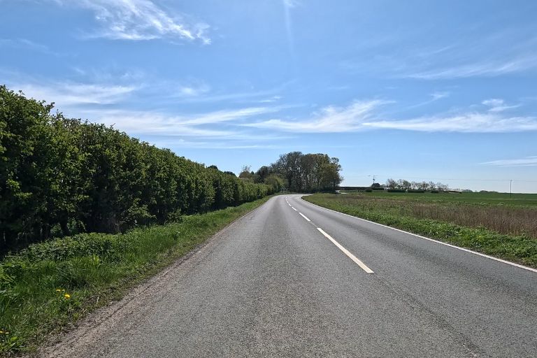 An empty straight road with bushes on one side and a green stretch of land on the other. The sky is blue with clouds dotted about.