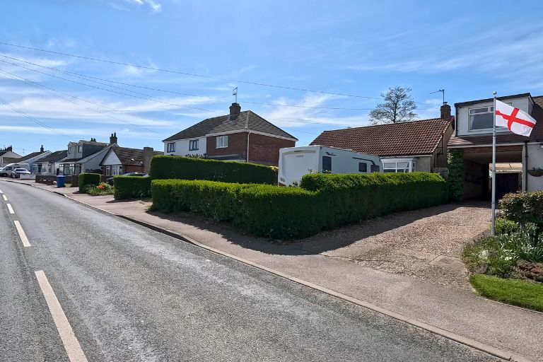 A typical looking British suburban road with houses on one side, with green hedges outlining the boundaries between each house. In the foreground is the England flag, white cross on a white background.
